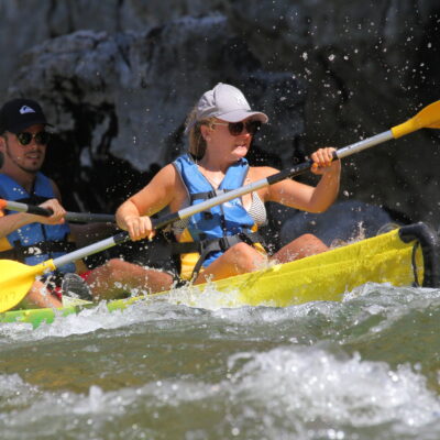 un canoe jaune et vert passe un rapide sur la rivière Ardèche, un homme et une femme. lui porte une casquette noire quiksilver et elle une casquette grise, ils ont tous les 2 une pagaie double jaune
