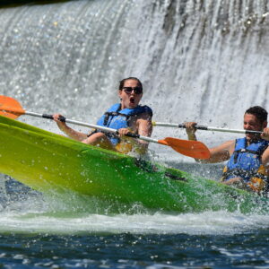 une femme et un homme à la sortie d'un tobboggan ou glissière sur l'ardèche à Sampzon, la femme est surprise par l'eau, le garçon sourit, derrière on aperçoit une chute, un bateau canoe jaune et vert une pagaie orang et une jaune