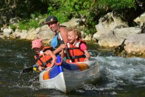 1 adulte guide dans un canoe ouvert vogueur bleu, orange et rouge et blanc, les enfants rigolent ils ont des gilets de sauvetages oranges