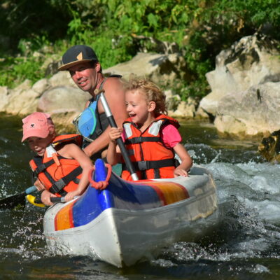 1 adulte guide dans un canoe ouvert vogueur bleu, orange et rouge et blanc, les enfants rigolent ils ont des gilets de sauvetages oranges