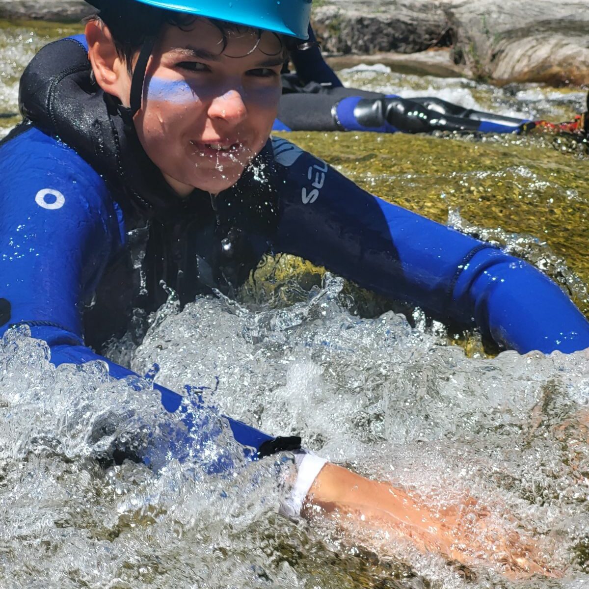 un adolescent dans une combinaison néopréne noire et bleu avec un casque bleu petzl, il est assis dans l'eau il fait du canyoning, il a des traces de crème solaire bleu sur les joues