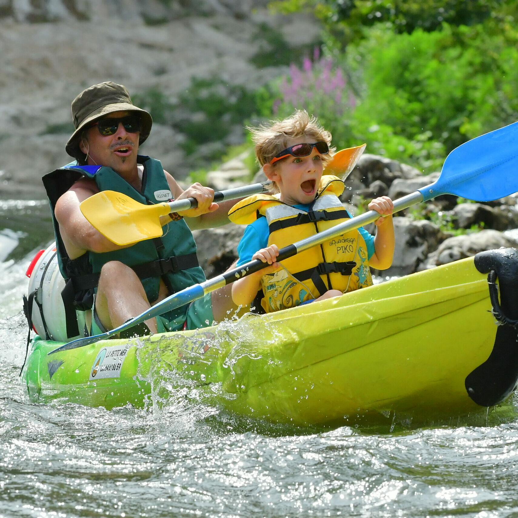 le papa et son fils en canoë de location en Ardèche jaune et vert, l'enfant à l'avant porte un gilet de sauvetage jaune aquadesign, il porte des lunettes de soleil, la papa porte un gilet de sauvetage vert, il porte un bob vert kaki et des lunettes de soleil, il passe un rapide le petit garçon crit ah. Il fond du canoe sur la rivière ardèche, il passe un rapide, on aperçoit l'eau et les caillous derrière eux