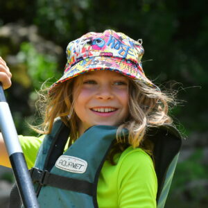 Un enfant avec un bob et un gilet de sauvetage sur un canoë tient une pagaie et sourit