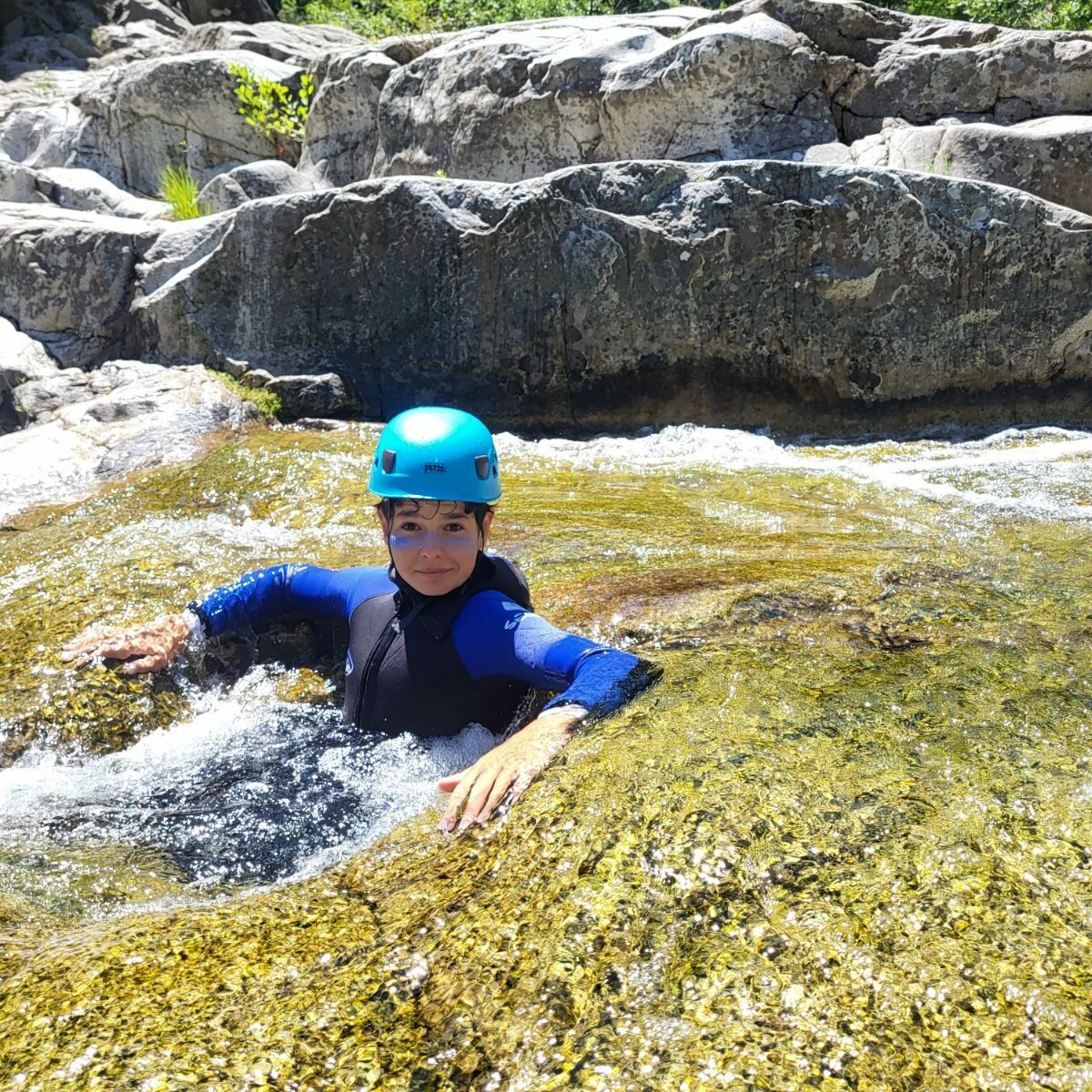 un adolescent est assis dans une marmitte d'eau, il porte une caombinaiosn néoprène noire et bleue sealand et un casque bleu petzl, il a de la crème solaire bleu sur le visage, il sourit