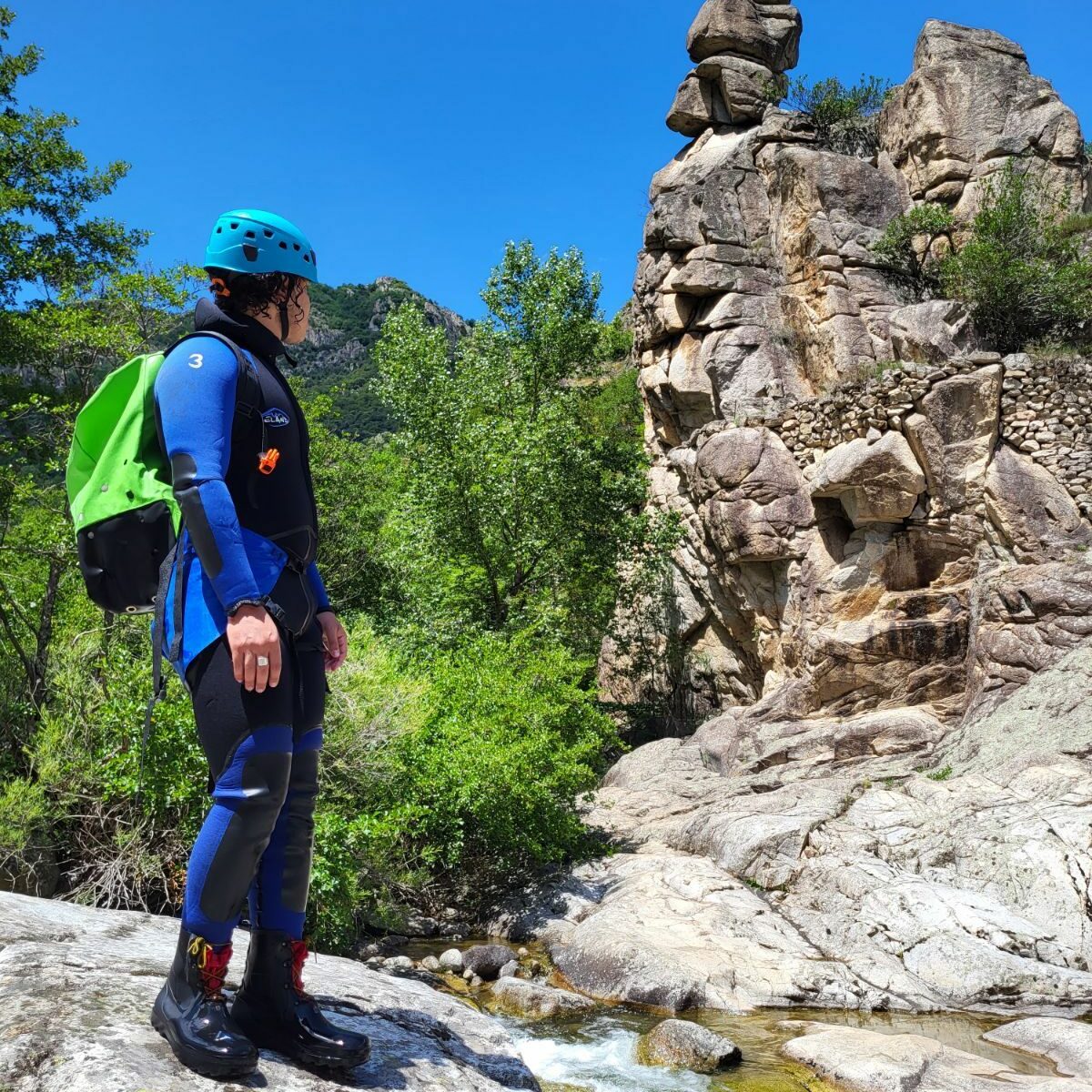 un homme est debout et regarde un rocher en granite, il porte une combinaison noire et bleue et un sac de canyon vert petz ainsi qu'un casque bleu petzl