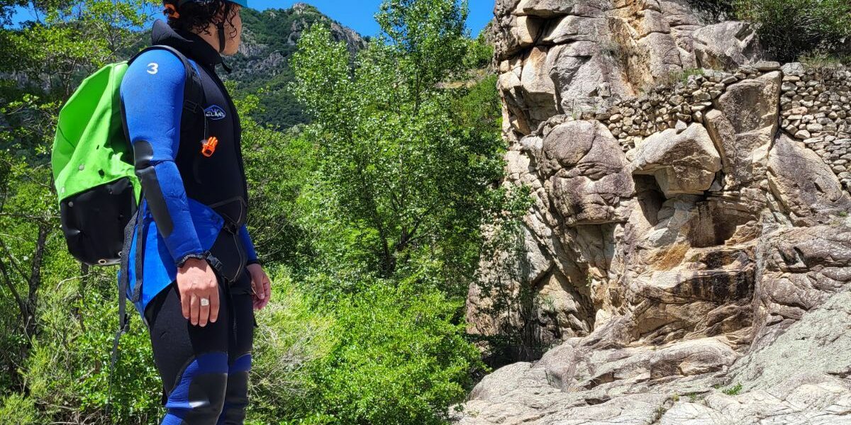 un homme est debout et regarde un rocher en granite, il porte une combinaison noire et bleue et un sac de canyon vert petz ainsi qu'un casque bleu petzl