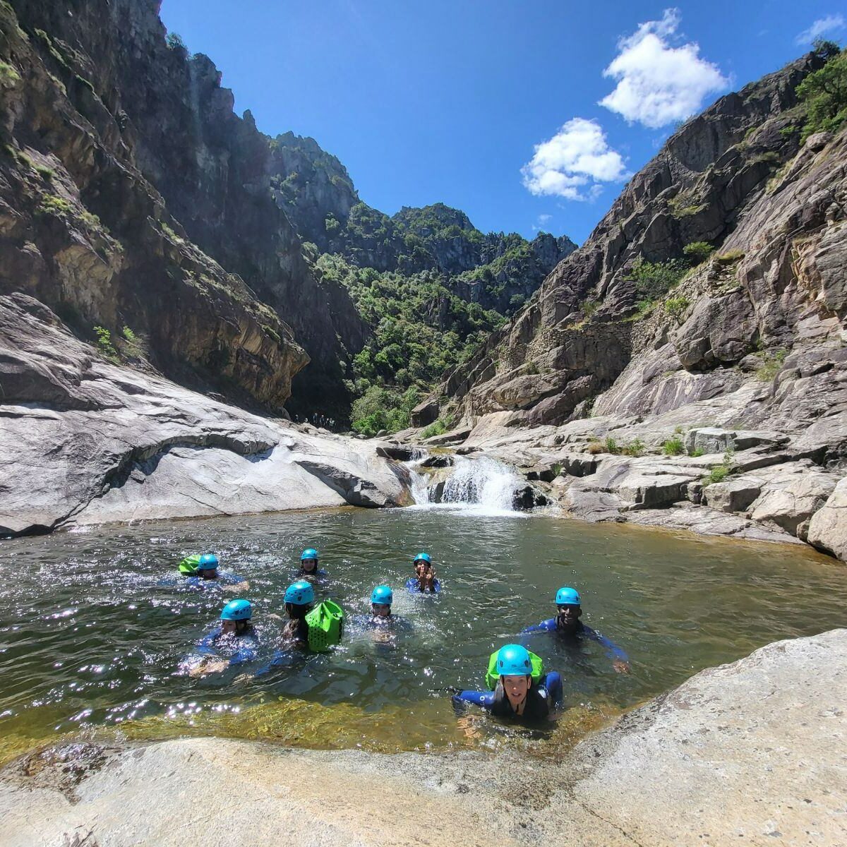 8 jeunes sont dans une vasque d'eau sur le canyon du Chassezac, ils portent des combinaisons néoprène sealand noires et bleues, des sacs de canyon verts petzl, on voit des rochers de graites et de l'eau qui coule d'une petite cascade, le ciel est bleu avec 2 nuages blancs