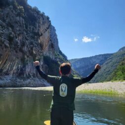 Un enfant est debout de dos sur un paddle sur l'eau levant les 2 bras