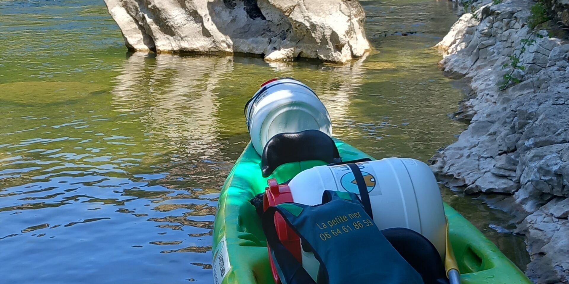 canoe bivouac un canoë double jaune et vert avec 2 bidons étanches, un gilet de sauvetage est posé sur le canoë aisni qu'une pagaie, l'eau est translucide, un rocher au milieu de la rivière.