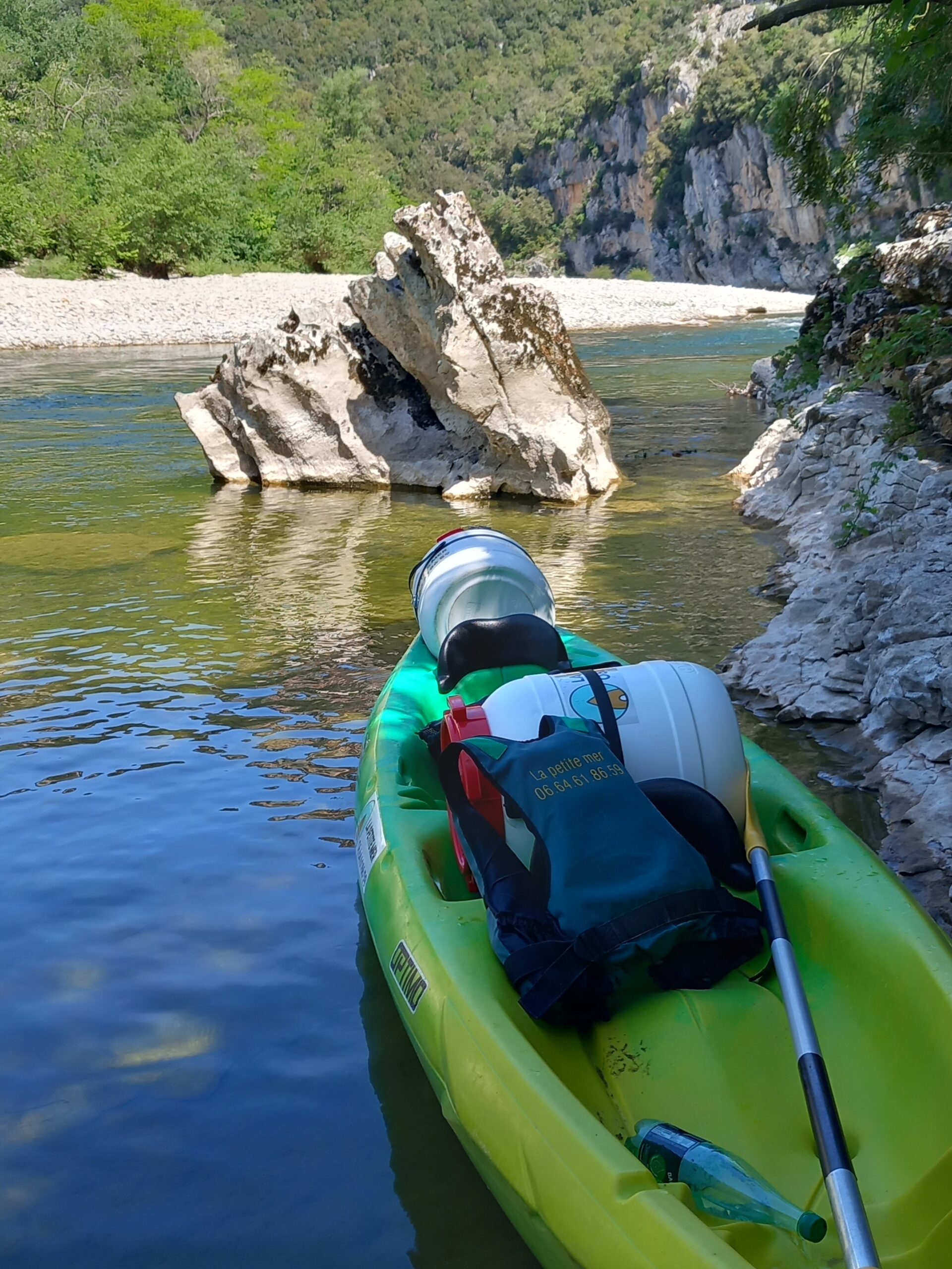 un canoë double jaune et vert avec 2 bidons étanches, un gilet de sauvetage est posé sur le canoë aisni qu'une pagaie, l'eau est translucide, un rocher au milieu de la rivière.