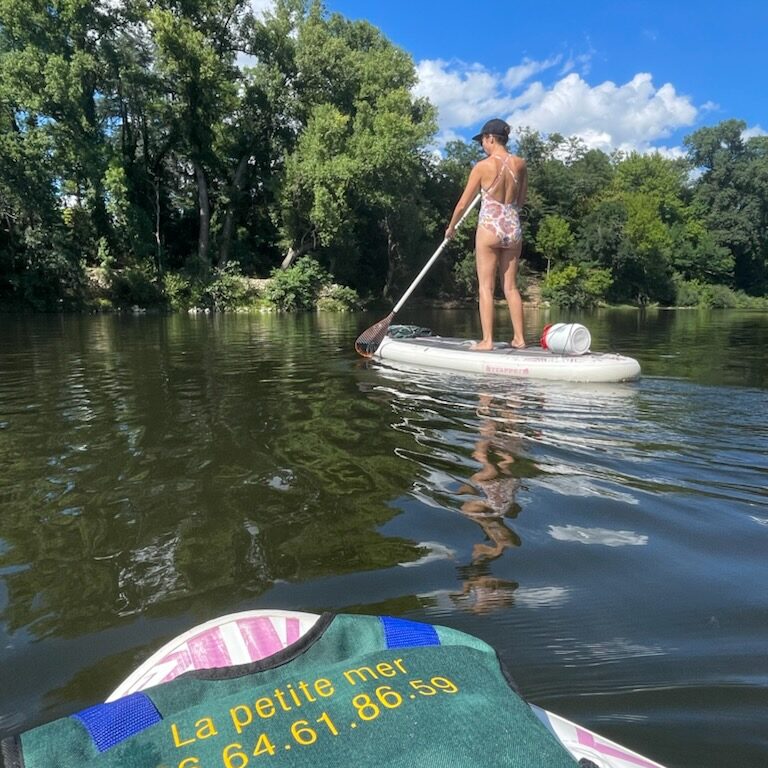 une jeune femme en maillot de bain à fleurs sur un paddle blanc avec une pagaie simple et un petit bidon étanche blanc sur l'arrière du paddle, elle porte une casquette noire. On aperçoit un bout de paddle b