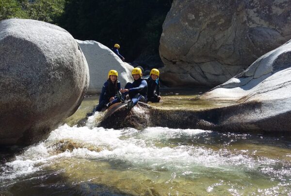 Three children in a canyon wearing black and blue Sealands wetsuits with yellow helmets. Behind them, we can see a high cliff. They are sitting in a pool, and the water is crystal clear.