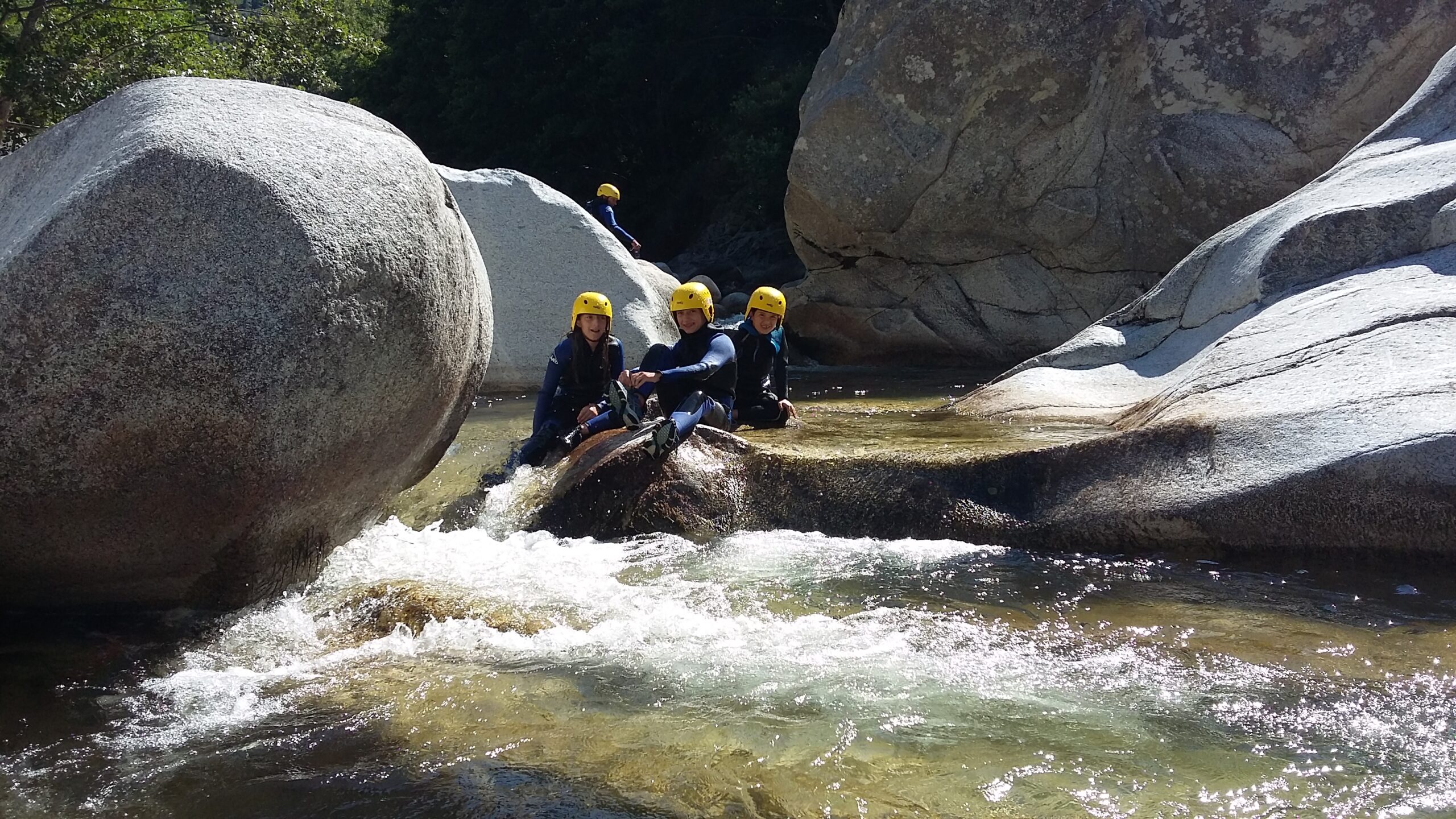 Three children in a canyon wearing black and blue Sealands wetsuits with yellow helmets. Behind them, we can see a high cliff. They are sitting in a pool, and the water is crystal clear.