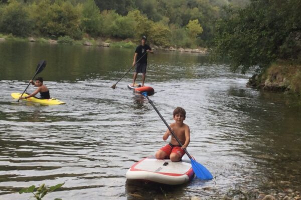 3 paddles 1 enfant à genou avec une pagaie simple, un adulte debout avec une pagaie dans l'eau, un kayak freestyle jackson jaune sur la plage de la base nautique de la petite mer sur la rivière Ardèche