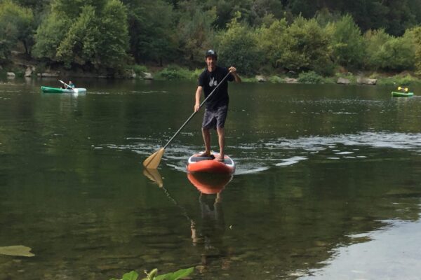 un homme avec un t-shirt noir et short de bain noir et une casquette sur un paddle à l'embarquement de la base nautique de la petite mer, derrière un bateau vert et une pagaie jaune et un autre blanc et vert clair