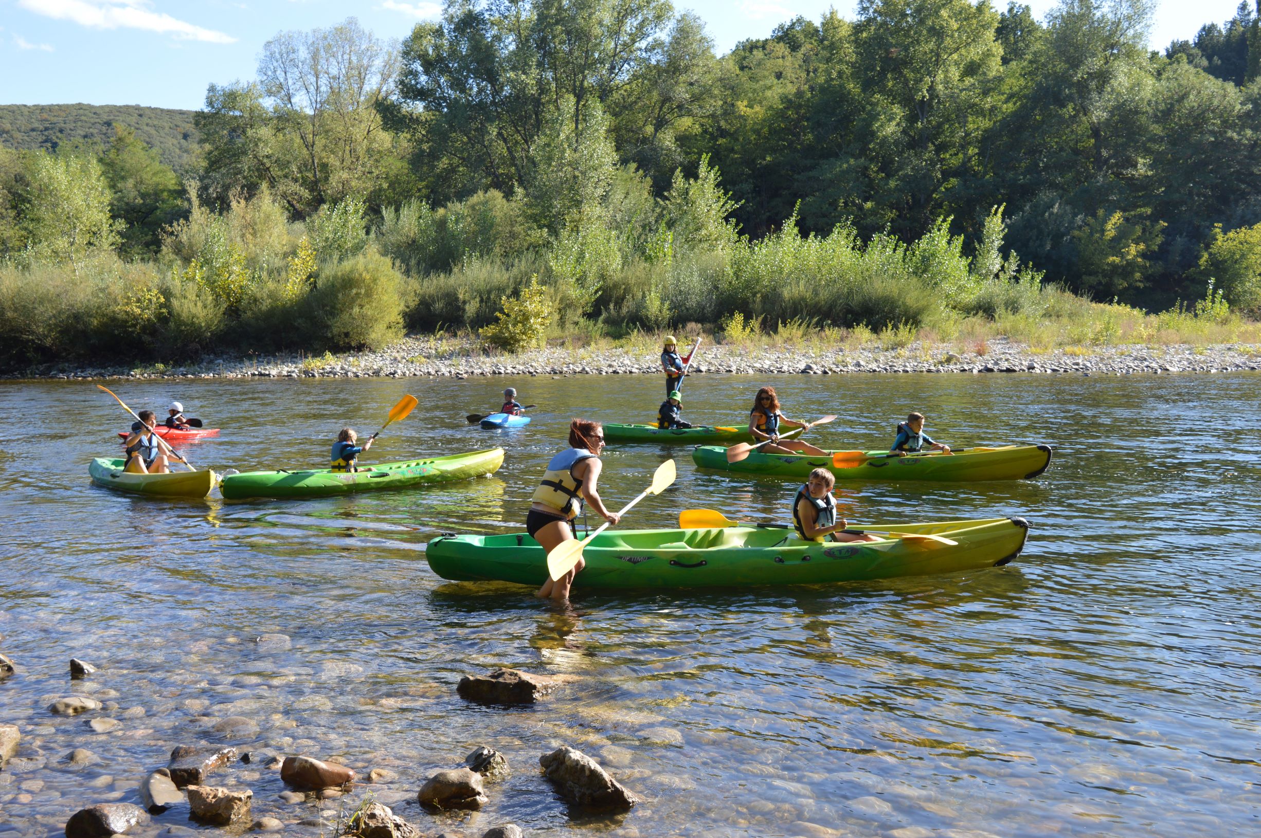 5 bateaux bi places jaunes et verts avec des femmes et des enfants qui portent des maillots de bain et gilets de sauvetages jaunes et bleus, 2 bateaux de rivières , un bleu et un rouge. A l'embarquement de la base nautique de la petite mer