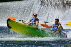 une femme et un homme à la sortie d'un tobboggan ou glissière sur l'ardèche à Sampzon, la femme est surprise par l'eau, le garçon sourit, derrière on aperçoit une chute, un bateau canoe jaune et vert une pagaie orang et une jaune