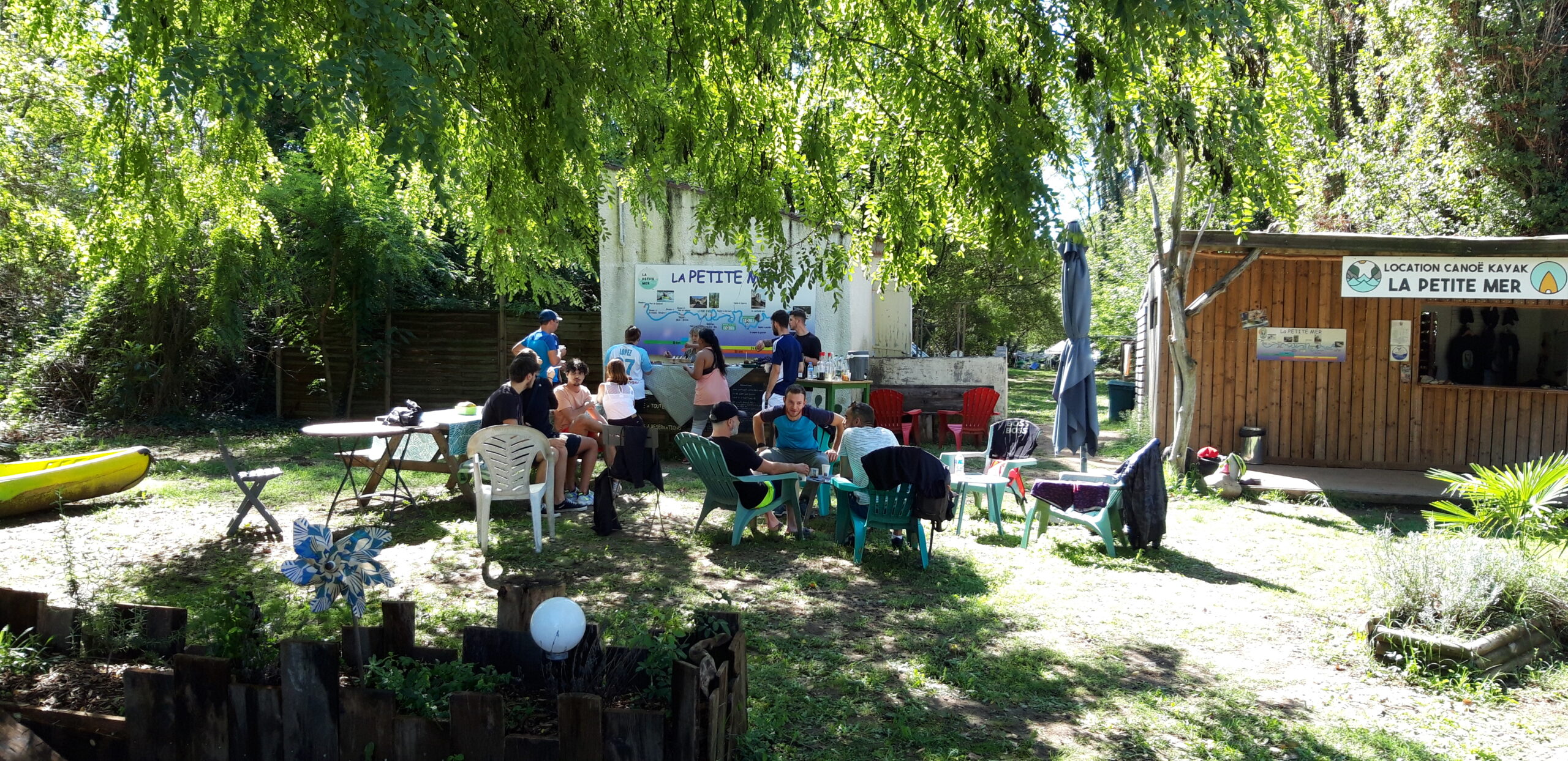Le terrain de la Petite Mer en Ardèche ombragé avec une réception, du matériel de canoë kayak et des gens assis sur des chaises