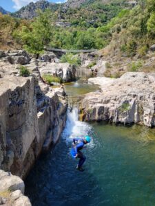 un homme en combinaison noire et bleu avec un casque bleu petzl saute dans la rivière derrière on aperçoit un pont de singz