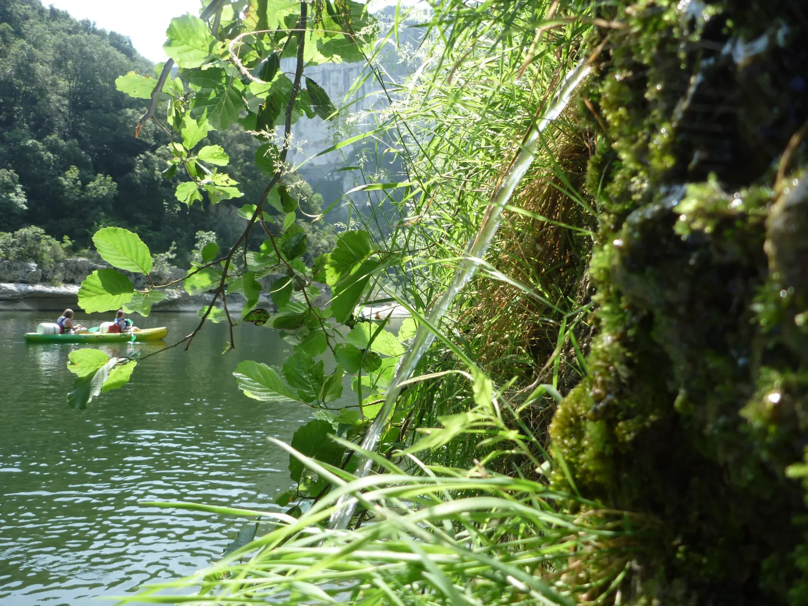 derrière des végétaux on aperçoit un bateau jaune et vert , il y a 2 adultes qui portent des gilets de sauvetages. deux bidons sont installés. ils naviguent sur l'Ardèche et se préparent à faire un bivouac. on voit les falaises et de la végétation autour d'eux